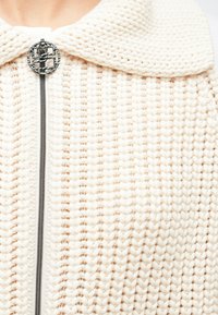 Kardigan dziany w kolorze ecru, z dużym kołnierzem, srebrnym zamkiem błyskawicznym i ozdobnym czarnym guzikiem o skręconym wzorze. Gruba faktura.