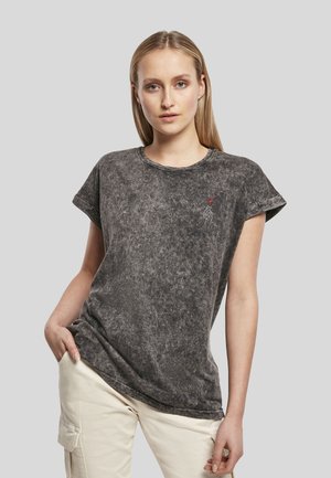 Jeune femme aux cheveux blonds raides portant un t-shirt gris effet délavé avec manches retroussées et un pantalon cargo beige, debout devant un fond uni.