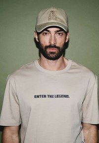 Beżowa koszulka z napisem "WEJDŹ DO LEGENDY." w ciemnoniebieskim kolorze, w połączeniu z jasnobeżową czapką zdobioną logo.