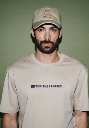 Beżowa koszulka z napisem "WEJDŹ DO LEGENDY." w ciemnoniebieskim kolorze, w połączeniu z jasnobeżową czapką zdobioną logo.