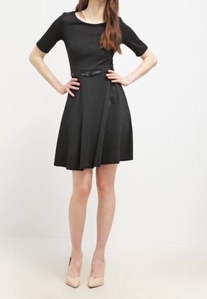 Femme portant une robe noire longueur genou avec des manches courtes et une fine ceinture, associée à des chaussures à talons beiges, debout avec les mains sur les hanches.