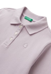 Helllila Polohemd mit drei weißen Knöpfen, einem umgeschlagenen Kragen und einem kleinen gestickten Logo auf der Brust mit einem grünen Markenetikett innen.