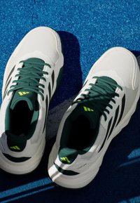 Baskets blanches avec des lacets et des accents verts, présentant une surface texturée et des rayures noires. Semelle antidérapante avec un talon renforcé.