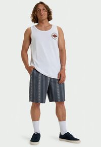 Man with curly hair wearing a white sleeveless shirt with a logo, striped gray shorts, white socks, and black sneakers, standing with hands in pockets.