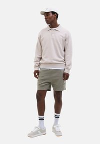 Lichtbeige polo sweatshirt, olijfgroene shorts en witte sneakers; met gestreepte sokken en een witte pet. Eenvoudig, ontspannen pasvorm ontwerp.