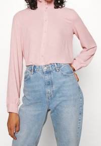 Blouse longue à manches longues rose clair avec un col haut et des détails de boutons, associée à un jean en denim bleu clair taille haute. Texture lisse.