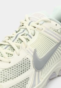Izbliza prikaz svijetle Nike tenisice s mrežastim panelima, rebrastim detaljima sa strane i sivoj Nike swoosh oznaci na boku.