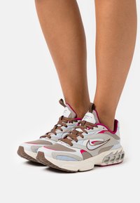 Grå och vita sneakers med rosa accenter och bruna snören. Har en texturerad ovandel och en tjock sula med synlig luftkuddar.