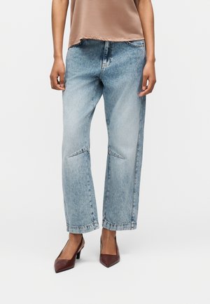 Άτομο που φοράει ανοιχτό μπλε τζιν παντελόνι με ίσια γραμμή και ραφή στο γόνατο, ένα μπεζ τοπ και καφέ γόβες με μυτερή μυτούλα, στέκεται μπροστά σε απλό φόντο.