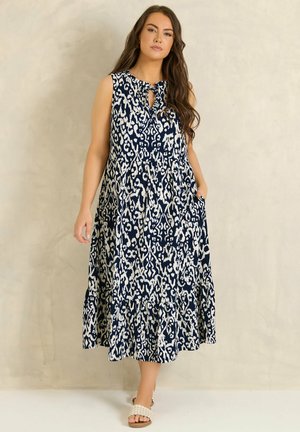 Frau trägt ärmelloses Midikleid in Marineblau mit weißem Muster und Volant am Saum, dazu Sandalen, steht vor einer beigen Wand.