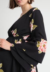 Černé květinové šaty s velkými růžovými a zelenými květy, dlouhými volánovými rukávy, výstřihem do V a elastickým pasem pro tvarování.