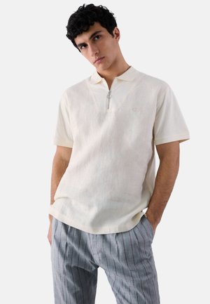 Giovane uomo che indossa una polo color panna a maniche corte con zip frontale e pantaloni grigi a righe, in piedi con le mani nelle tasche.