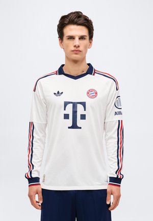 Junger Mann trägt ein weißes FC Bayern München Fußballtrikot mit langen Ärmeln, dunkelblauen und roten Streifen sowie Sponsorlogos und steht vor einem einfarbigen Hintergrund.