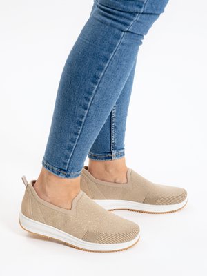 Person wearing beige slip-on sneakers with white soles and blue fitted jeans, standing on a plain white surface.