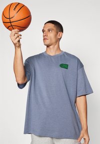 Gråblå oversized T-shirt med korte ærmer, prydet med et grønt grafik på brystet, sammen med grå sweatpants og en orange basketball.