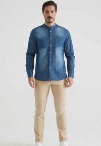 Homme portant une chemise en denim bleu à boutons et un pantalon beige, se tenant devant un fond blanc uni, faisant face.