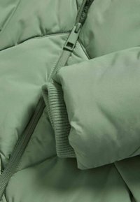 Grøn pufjakke med en glat tekstur, quiltet design og strikkede manchetter. Den har en lynlåslukning med en matchende lynlåsgreb.