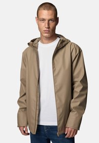 Beige wasserdichte Jacke mit Kapuze, Reißverschluss vorne und elastischen Bündchen, getragen über einem weißen T-Shirt, ergänzt durch blaue Jeans.