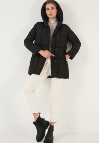 Zwarte pufferjas met capuchon, voorzien van een gestikt ontwerp, drukknoopsluiting en zijzakken. Model draagt witte broek en zwarte laarzen.
