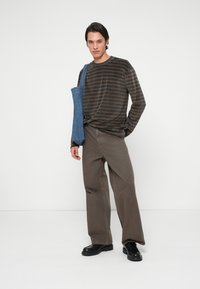 Νεαρός άνδρας φορεμένος με καφέ και μαύρες ρίγες μακρύ μανίκι πουκάμισο, χαλαρές καφέ παντελόνες, μαύρα παπούτσια, κρατώντας μια μπλε τσάντα χιαστί, στέκεται απέναντι σε λευκό τοίχο.