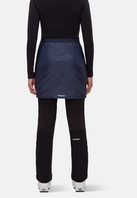 Marinblå isolerad kjol med quiltad struktur, kombinerad med svarta stretchbyxor och vit märkning. Buren med skidskor.