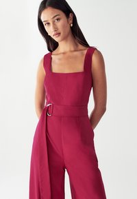 Ženska nosi rdež kombinezon brez rokavov s kvadratnim izrezom in ujemajočim se pasom iz blaga, ki ima srebrno okroglo zaponko.