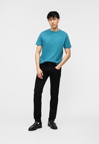 Νέος άνδρας που φοράει πετρόλ κοντομάνικο πουκάμισο, μαύρο παντελόνι και μαύρα μοκασίνια, στέκεται με το ένα χέρι στην τσέπη μπροστά από απλό φόντο.