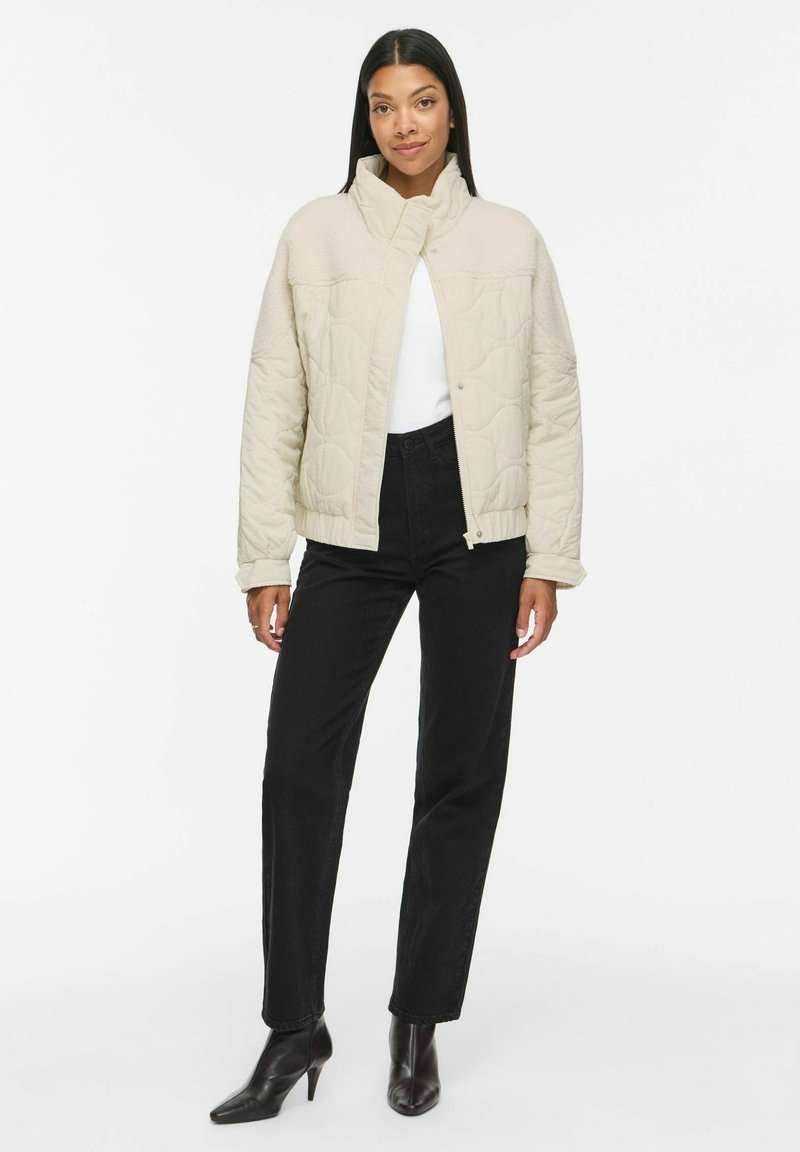 Femme debout portant une veste matelassée crème, une chemise blanche, un pantalon droit noir taille haute et des bottines noires sur fond blanc.