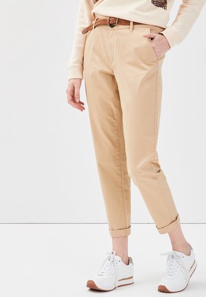 Personne portant un pantalon chino beige retroussé avec une ceinture tressée marron, un pull crème et des baskets blanches, debout devant un fond uni.