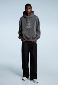 Grijze capuchontrui met een grafische print van een koffiekopje. Combineer met losse zwarte spijkerbroeken en lichtgekleurde sneakers. Gladde textuur.