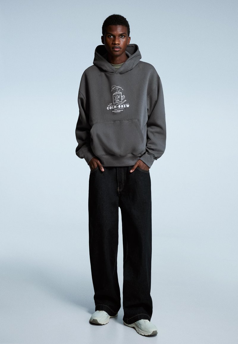 Grijze capuchontrui met een grafische print van een koffiekopje. Combineer met losse zwarte spijkerbroeken en lichtgekleurde sneakers. Gladde textuur.