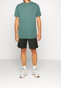 T-shirt en coton vert, col rond, manches courtes, associé à un short noir. Le mannequin porte des baskets blanches avec des accents rouges. Design minimaliste.
