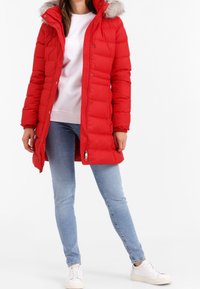 Röd vadderad jacka med pälsfodrad hjälm, hel dragkedja fram och tryckknappar. Kombinerad med en vit topp och ljusblå jeans. Vita sneakers.