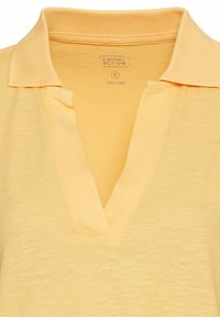Økologisk bomulds gul polo shirt med V-hals design og ribbet krave. Blød tekstur og subtil meleret mønster. Brandtag synlig indeni.