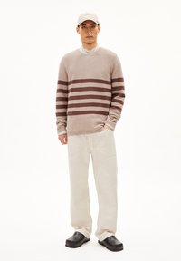 Maglione beige a righe con strisce orizzontali color maroon, abbinato a pantaloni chiari e zoccoli neri. Il modello si trova davanti a uno sfondo bianco.