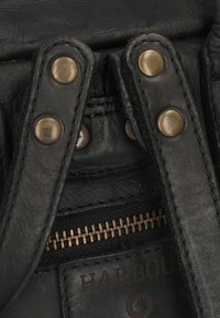 Primo piano di una borsa in pelle nera che mostra la superficie testurizzata, bottoni in ottone, e una cerniera metallica con fodera di tessuto scuro. "HARBOUR" impresso.