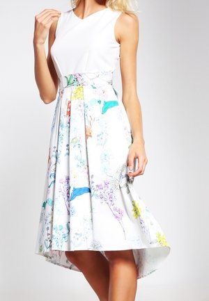 Femme portant une robe blanche sans manches avec un imprimé floral coloré, taille cintrée et ourlet asymétrique, debout devant un fond uni.
