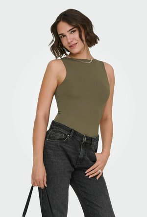 Kobieta o brązowych włosach do ramion, ubrana w oliwkowy, bezrękawnik i czarne dżinsy, trzyma pasek czarnej torebki na studiu.