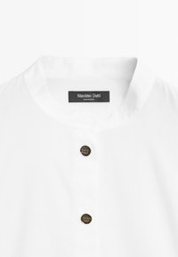 Chemise blanche avec un col rond et deux boutons décoratifs noirs et dorés sur le devant sous le col.