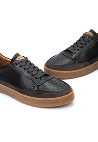 Sneaker in pelle nera con design perforato sulla punta, dettagli testurizzati e suola in gomma marrone. Presenta una chiusura con lacci e il logo del marchio.