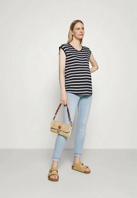 Mulher vestindo uma blusa sem mangas preta e branca de riscas, jeans skinny azul claro, sandálias bege, segurando uma bolsa de palha com alça castanha.