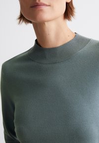 Grönstickad tröja med hög, ribbad halsringning. Slät textur, åtsittande design och långa ärmar. Enkel, ren estetik.