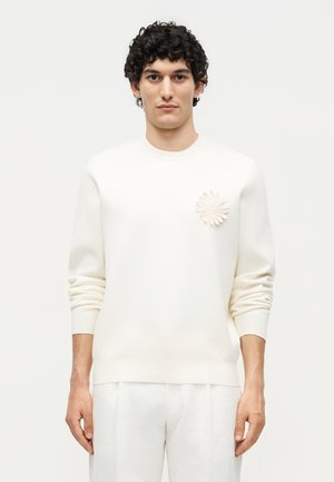 Mand med krøllet mørkt hår bærer en off-white sweater med en tekstureret blomsterbrosje på brystet og matchende off-white bukser.