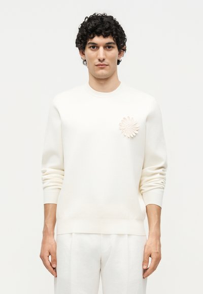 Homme aux cheveux bouclés foncés portant un pull écru avec une broche florale texturée sur la poitrine et un pantalon écru assorti.