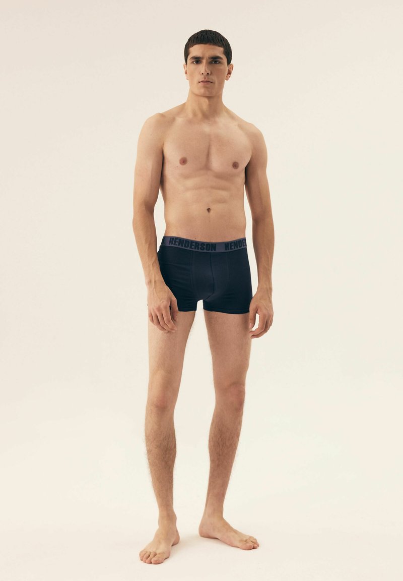 Czarne bokserki o gładkiej fakturze i szerokim, elastycznym pasku z napisem "HENDERSON" w kontrastowym kolorze. Model stoi boso.