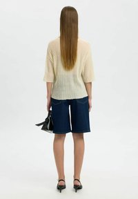 Cremfarbener, gerippter Strickpullover mit 3/4-Ärmeln, kombiniert mit dunklen Jeansshorts. Eine schwarze Handtasche und High Heels vervollständigen das Outfit. Sauberes, minimalistisches Design.