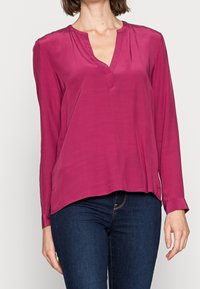 Blouse à manches longues en soie magenta vibrante avec un col en V et une coupe décontractée. Présente des plis subtils au niveau de l'encolure et un ourlet arrondi.
