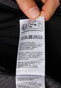Skötselråd på en svart textil, med flera språk. Texten inkluderar materialinformation: 94% polyester, 6% spandex, skötselråd.