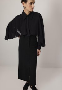 Black sheer blouse with a structured collar and flared sleeves, paired with a fitted black skirt featuring a front slit. Textured fabrics.