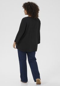Black long-sleeve top with a relaxed fit, featuring a curved hem and lightweight texture. Paired with blue wide-leg jeans and beige footwear.